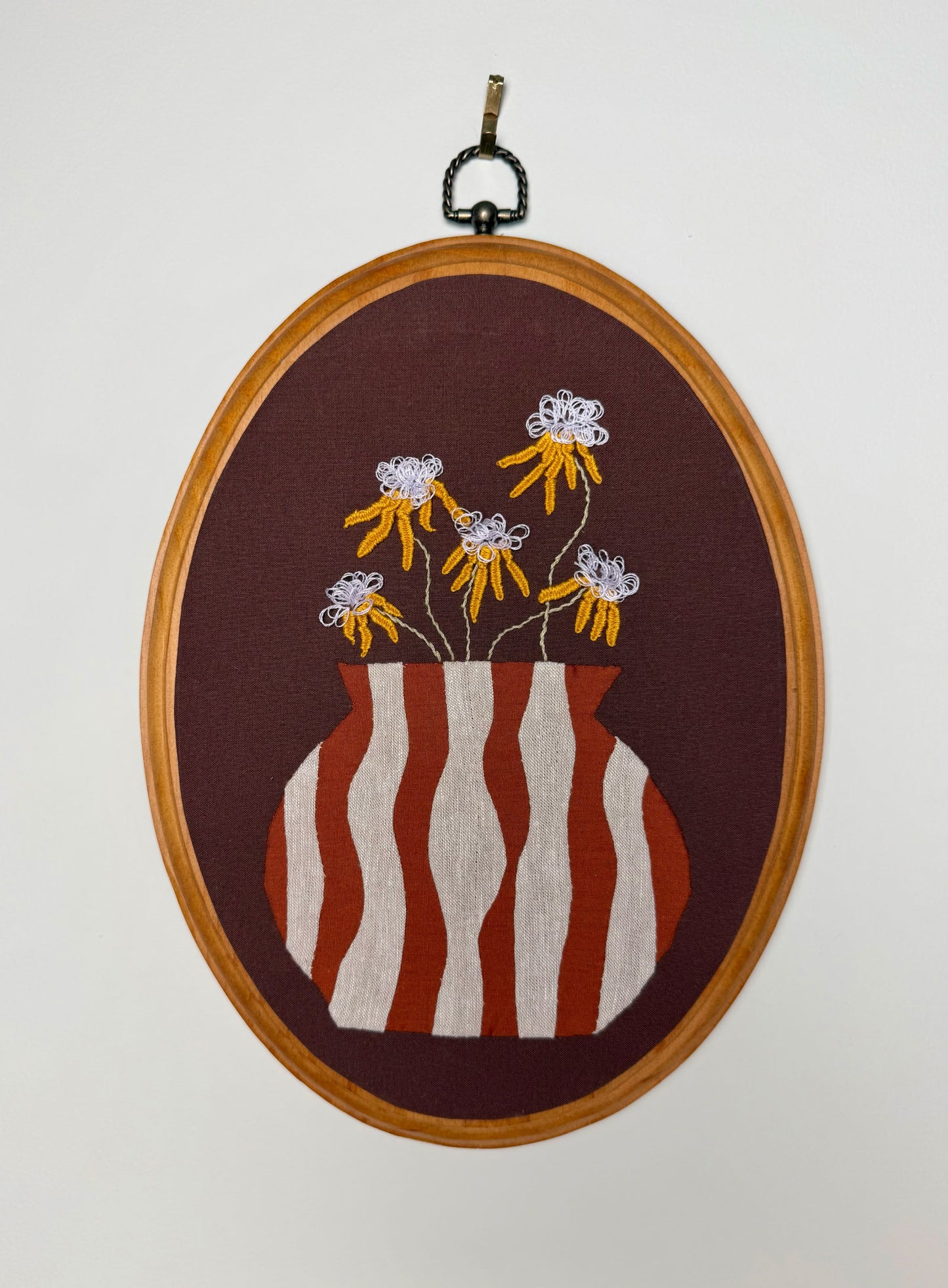 A wood oval framed embroidery featuring a striped appliqué vase with embroidered yellow flowers on a brown background, hanging against a plain light wall.
