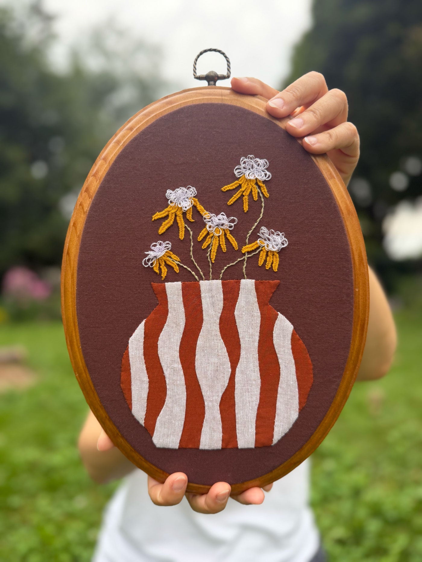 A person holds an oval wooden frame featuring a brown fabric background with a striped terracotta and white vase appliqué and embroidered yellow flowers.