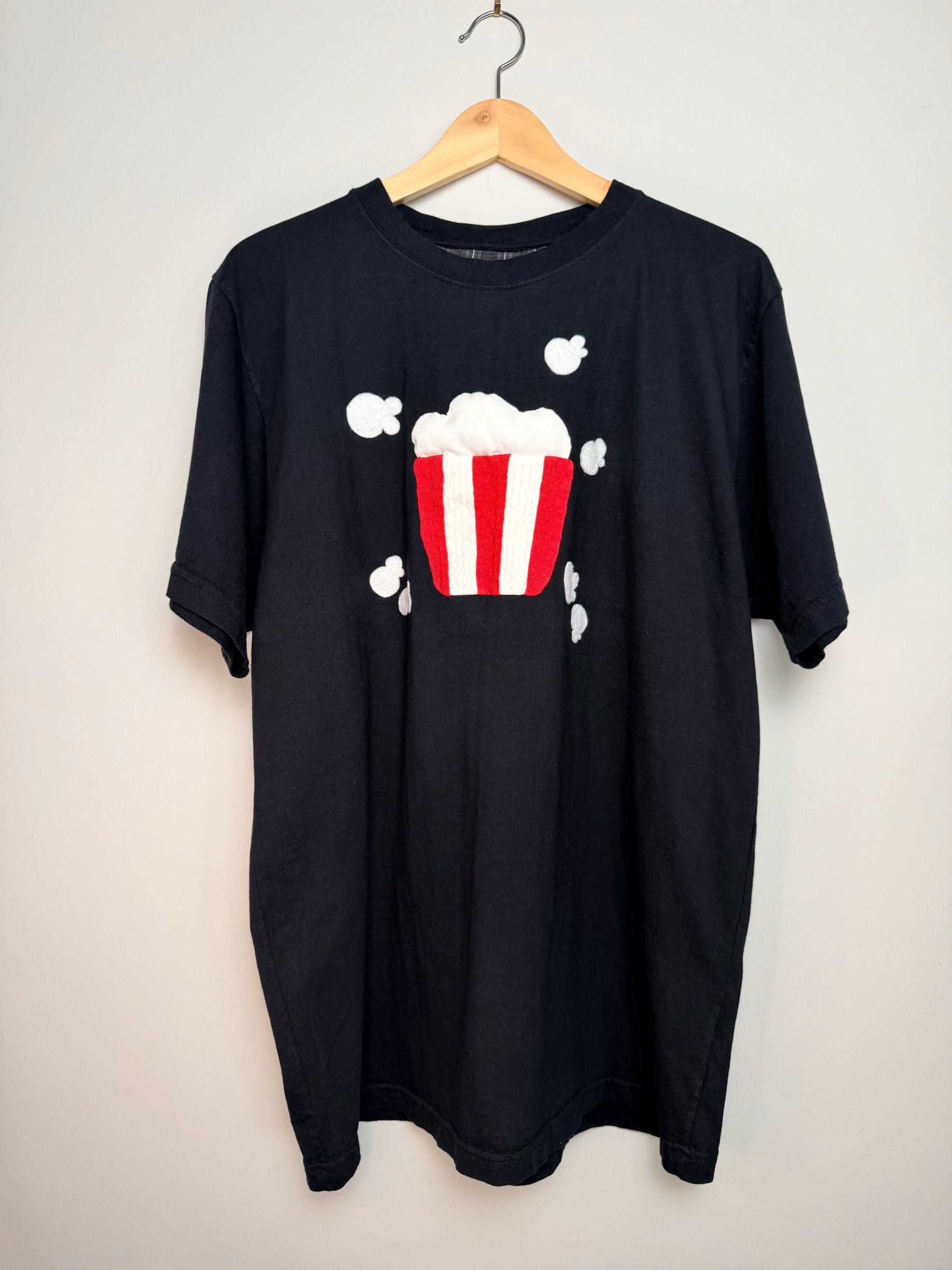 Black t-shirt with a popcorn appliqué design on a hanger against a grey background