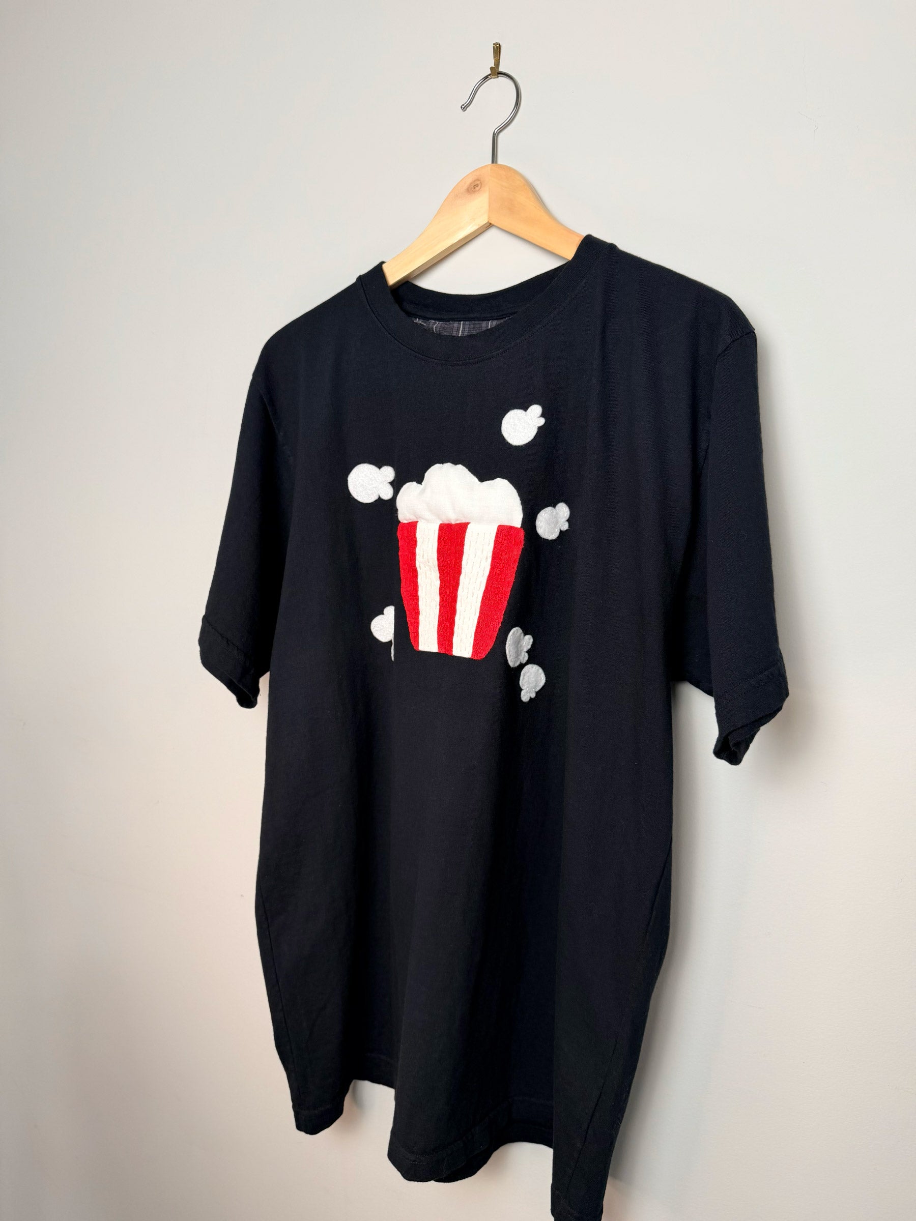 Black t-shirt with a popcorn appliqué design on a hanger against a grey background