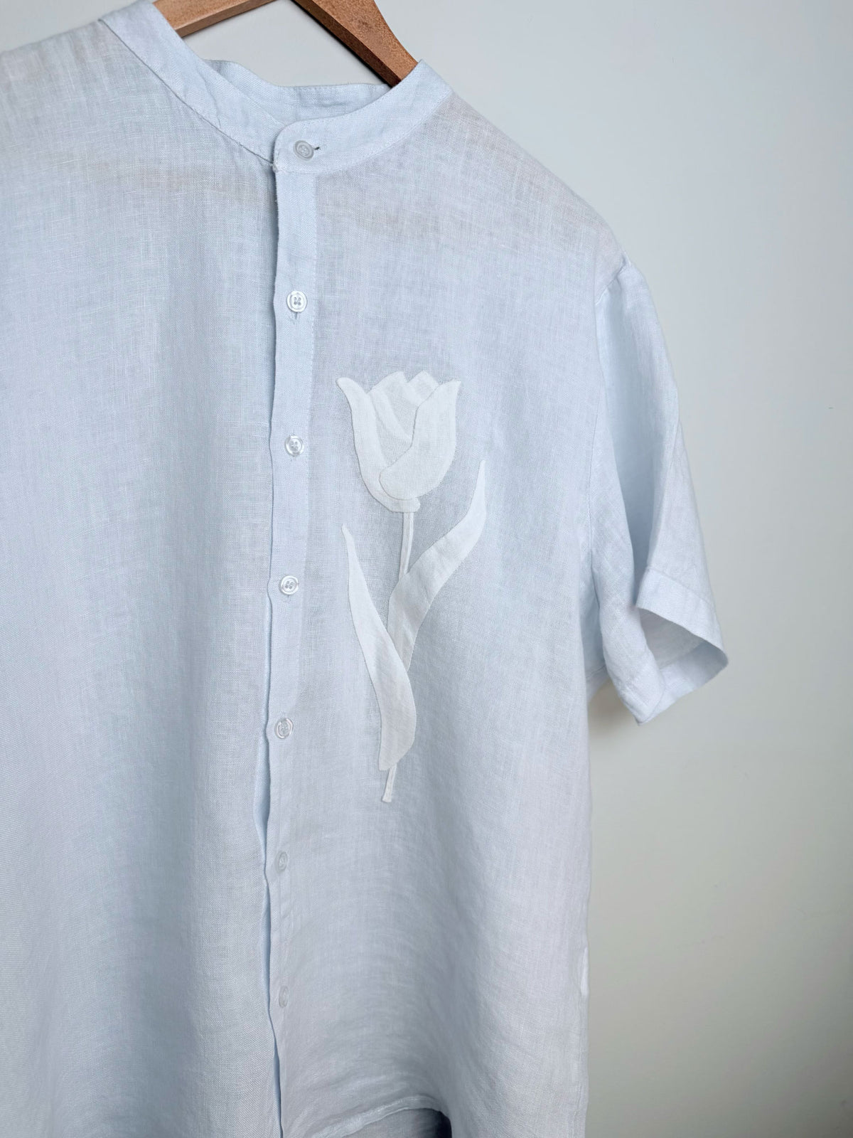 Light blue linen shirt with a off white tulip appliqué design on a hanger against a grey background