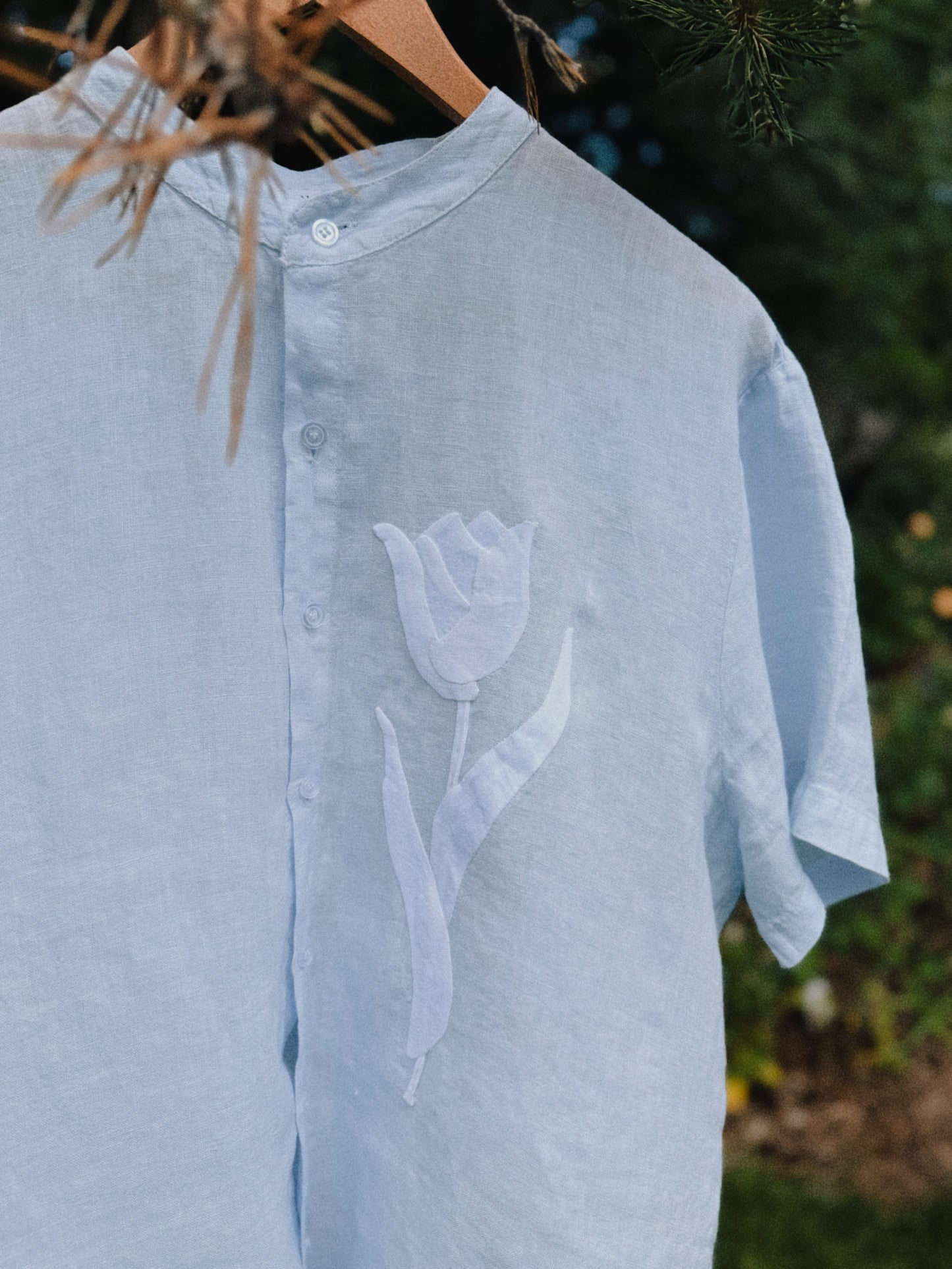 Light blue linen shirt with off white tulip appliqué design on a blurred natural background