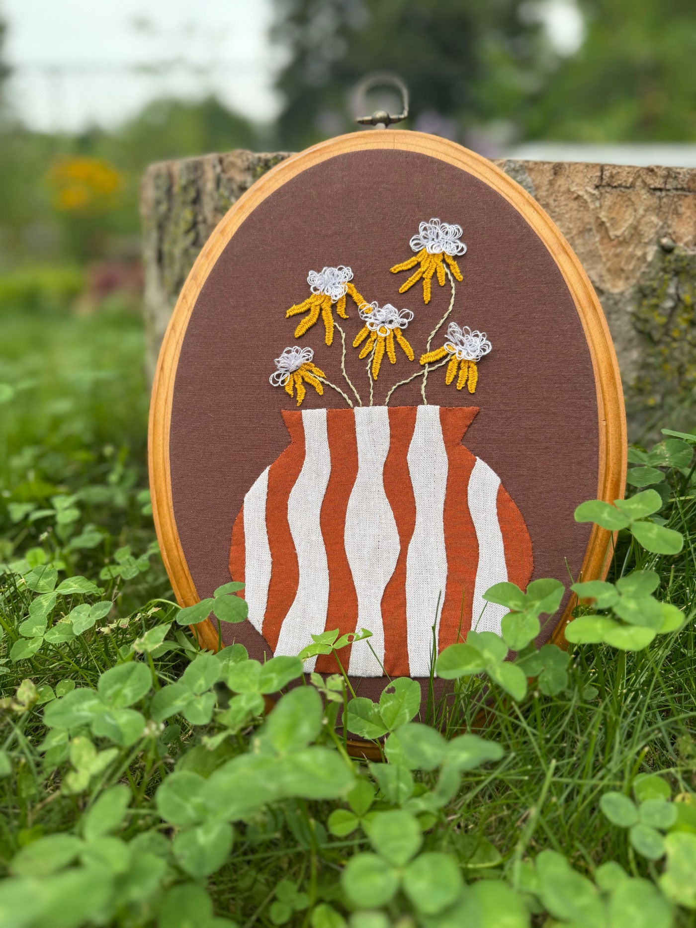 A wood oval frame featuring an embroidered floral design with an appliqué vase, displayed outdoors on green grass in front of a tree trunk.