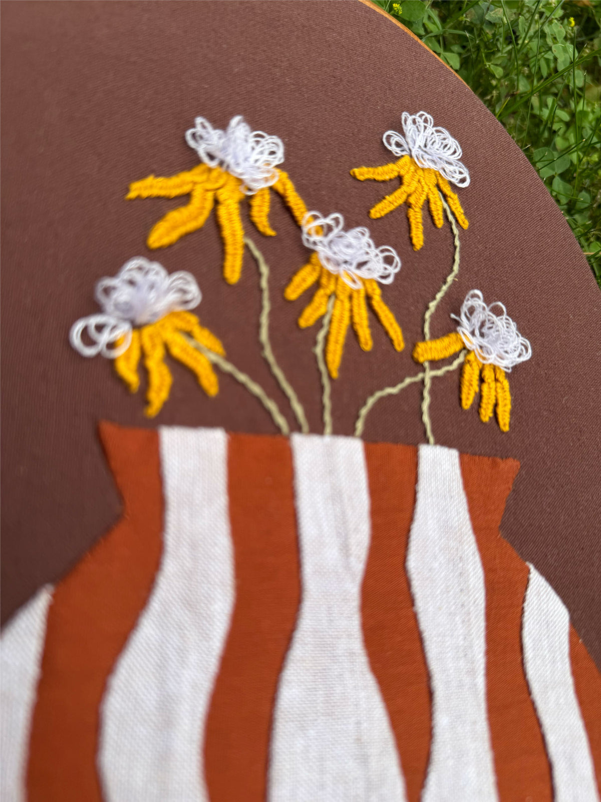 A close-up of a decorative textile wall hanging featuring embroidered flowers with a striped appliqué vase motif on a brown background.