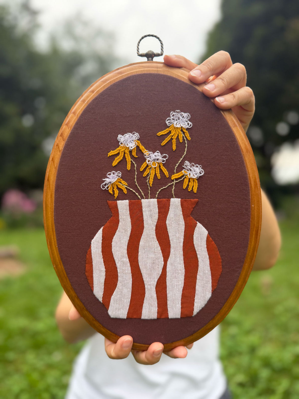 A person holds an oval wooden frame featuring a brown fabric background with a striped terracotta and white vase appliqué and embroidered yellow flowers.