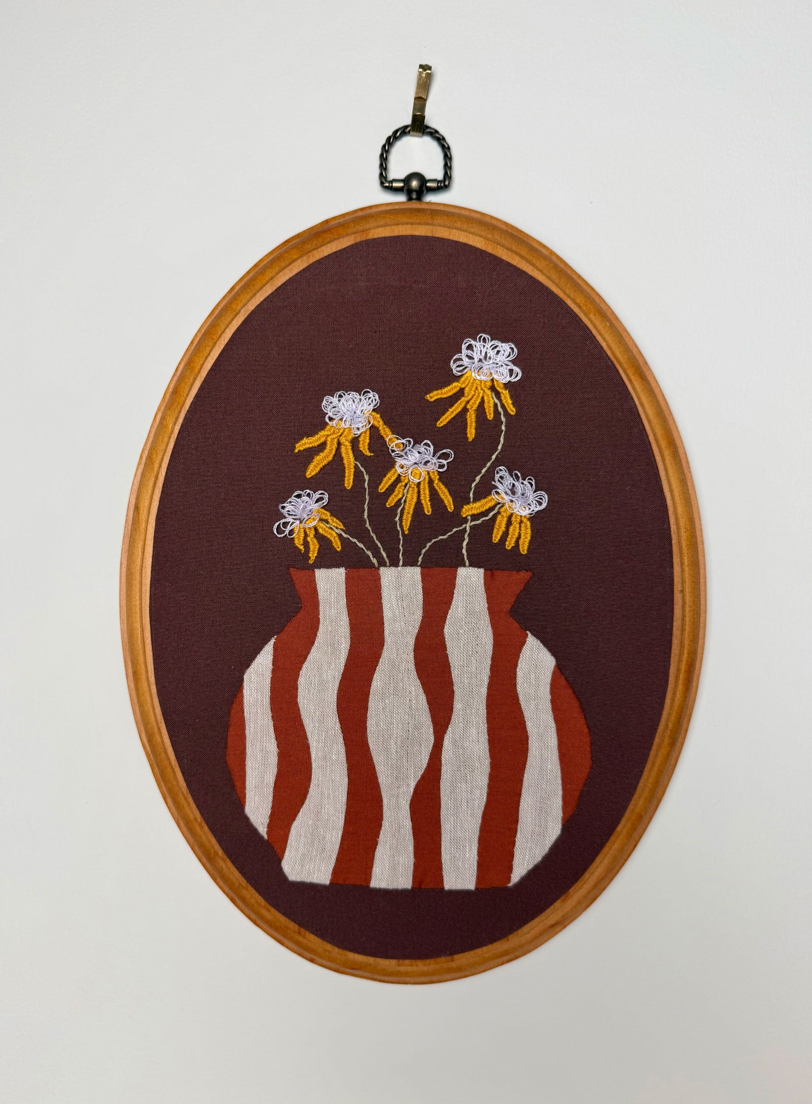 A wood oval framed embroidery featuring a striped appliqué vase with embroidered yellow flowers on a brown background, hanging against a plain light wall.
