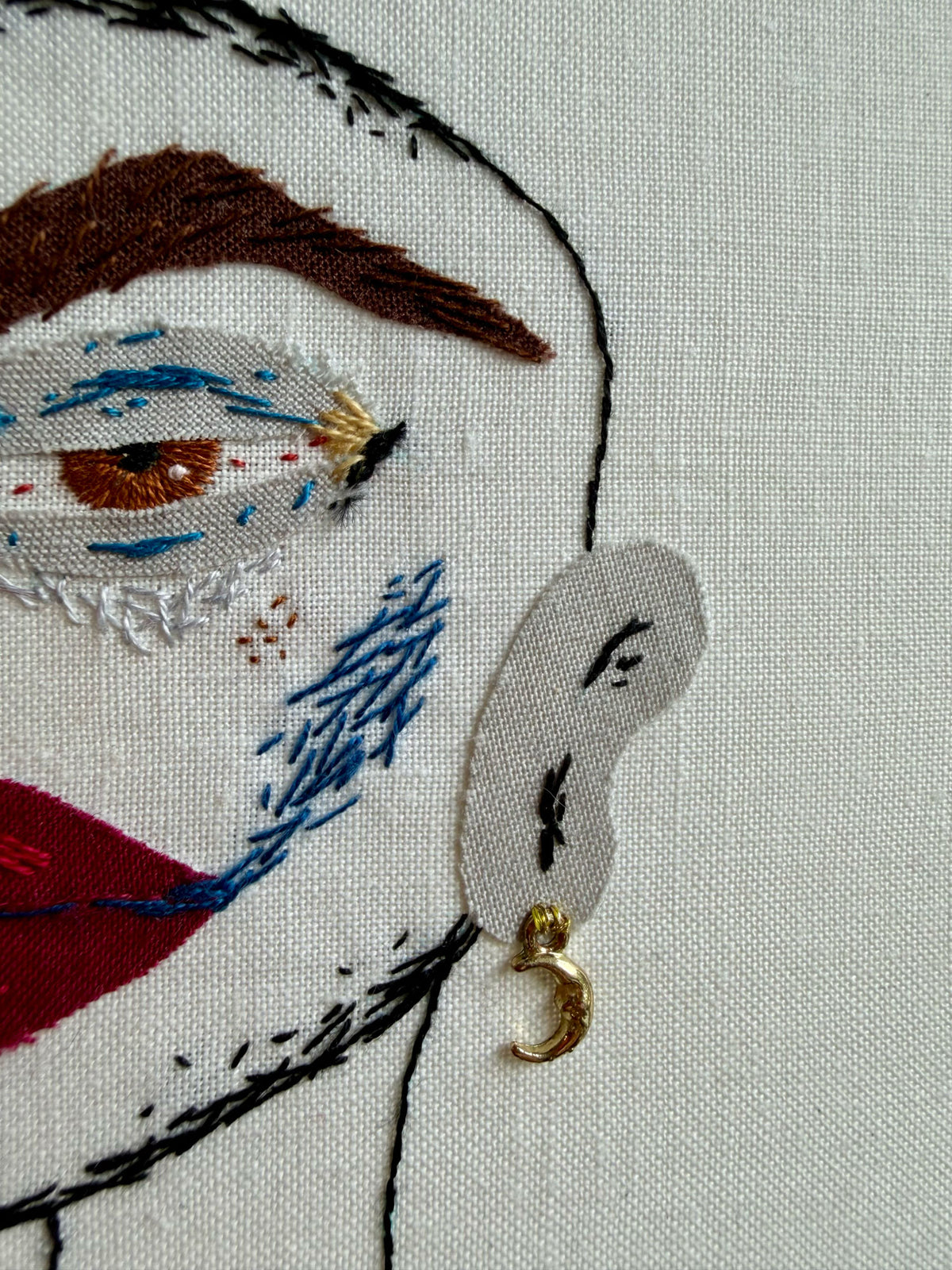 Close-up of an embroidered textile portrait showing part of a face with brown hair, blue and white eye details, red lips, and a small gold crescent earring attached near the ear area on off-white fabric.