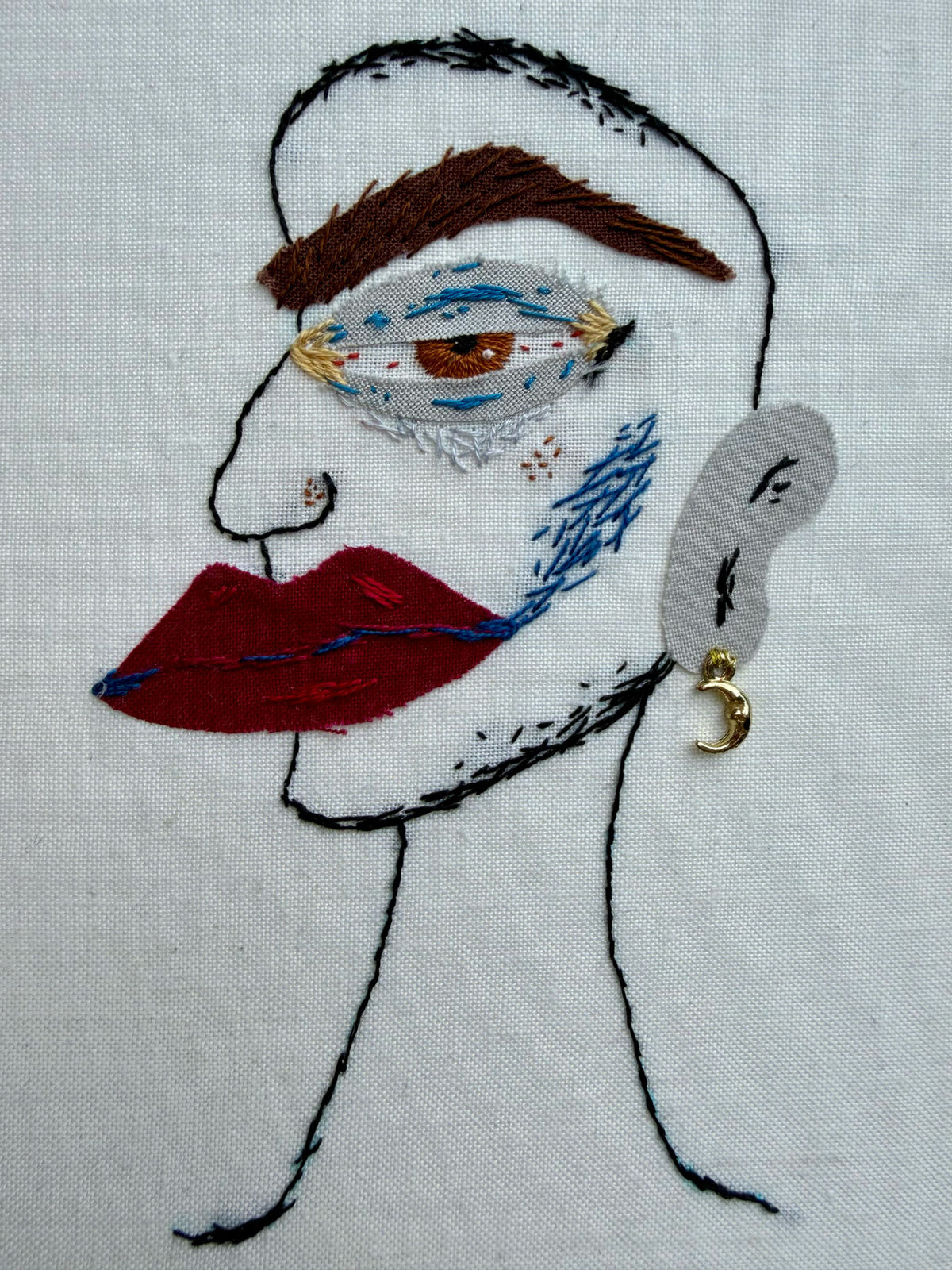 A hand-embroidered face on off white fabric, featuring a brown eyebrow, a stylized eye with blue and white detailing, red lips, a grey ear, and a small gold crescent moon earring.