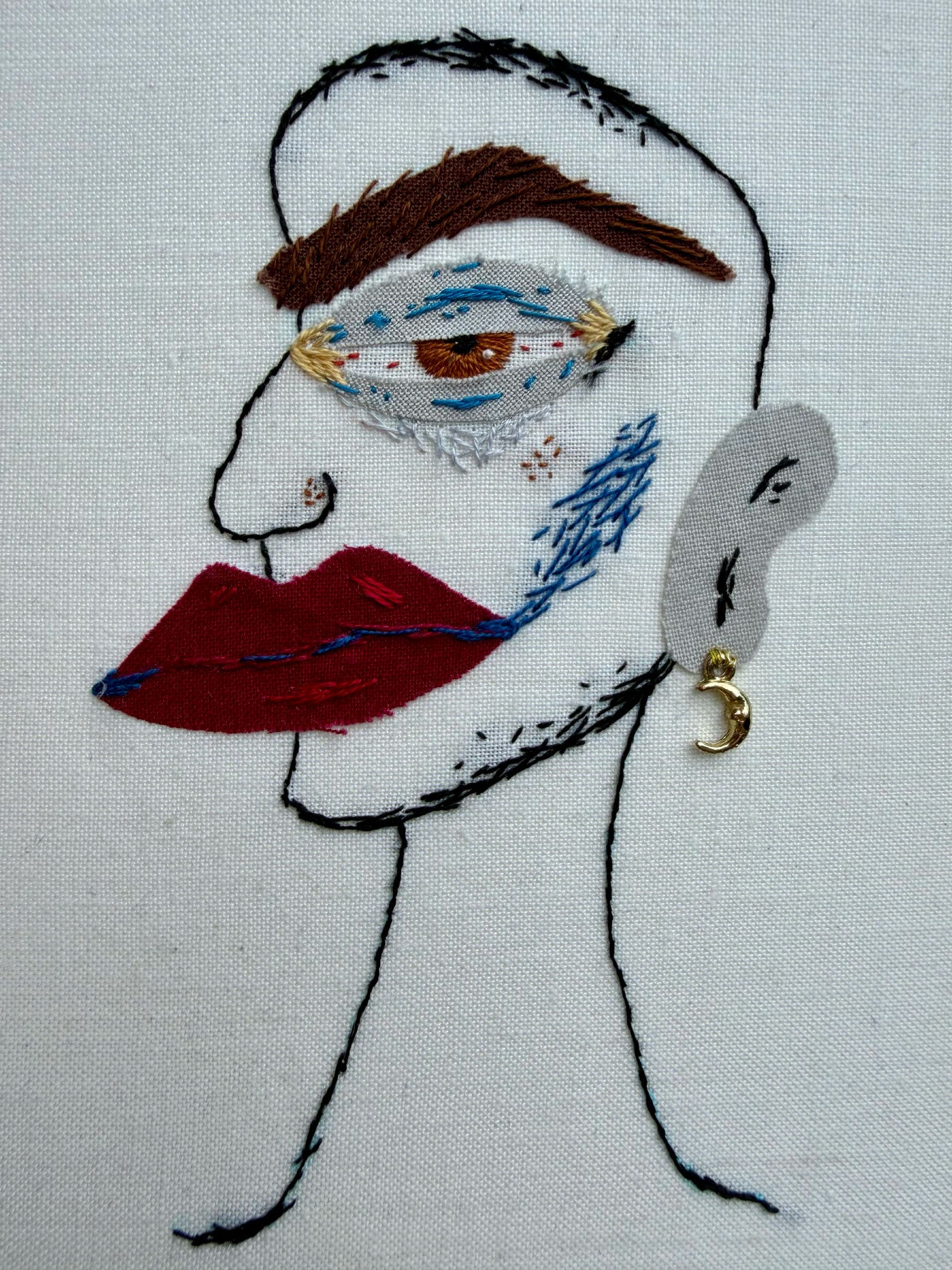 A hand-embroidered face on off white fabric, featuring a brown eyebrow, a stylized eye with blue and white detailing, red lips, a grey ear, and a small gold crescent moon earring.