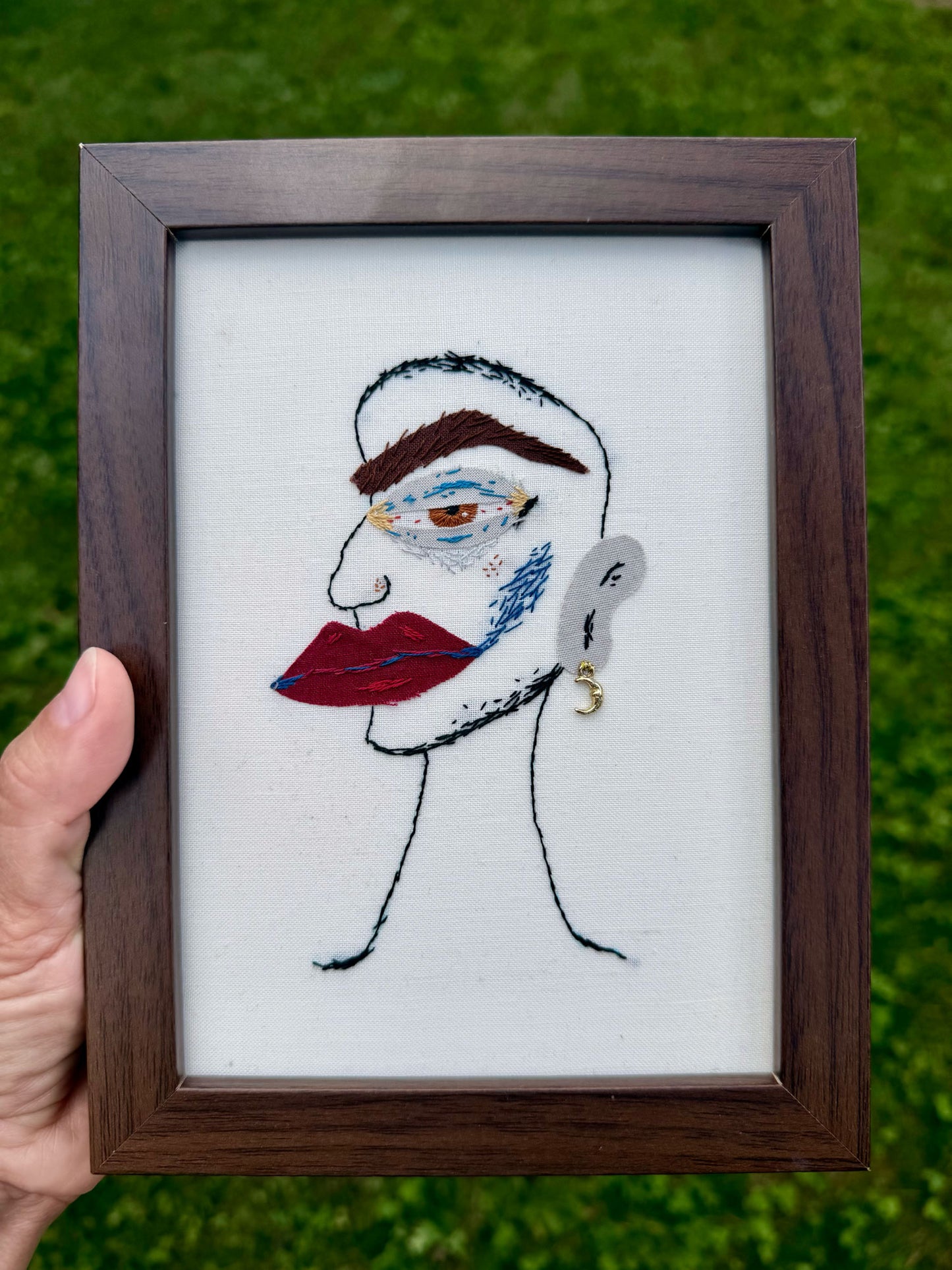 A square framed artwork featuring a embroidered face with red lips, blue and brown details, and a small gold earring on a off white fabric background. The frame is dark brown wood, set against a blurred green grass background.