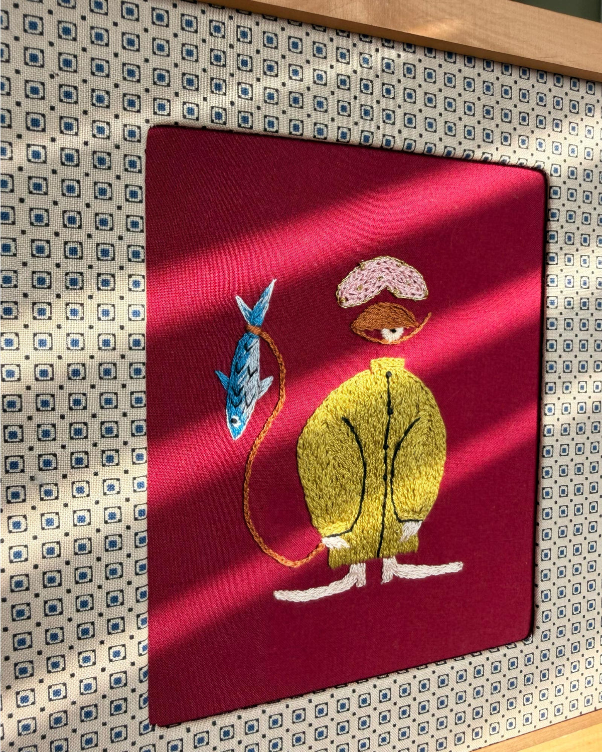 Embroidery of a character with a fish on a red background, assembled in a wood frame.