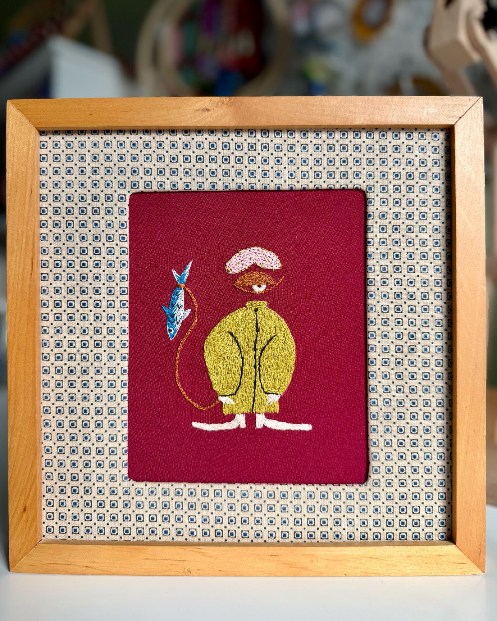 Embroidered picture of a character with a fish on a red background, framed in a wooden frame with a patterned mat.