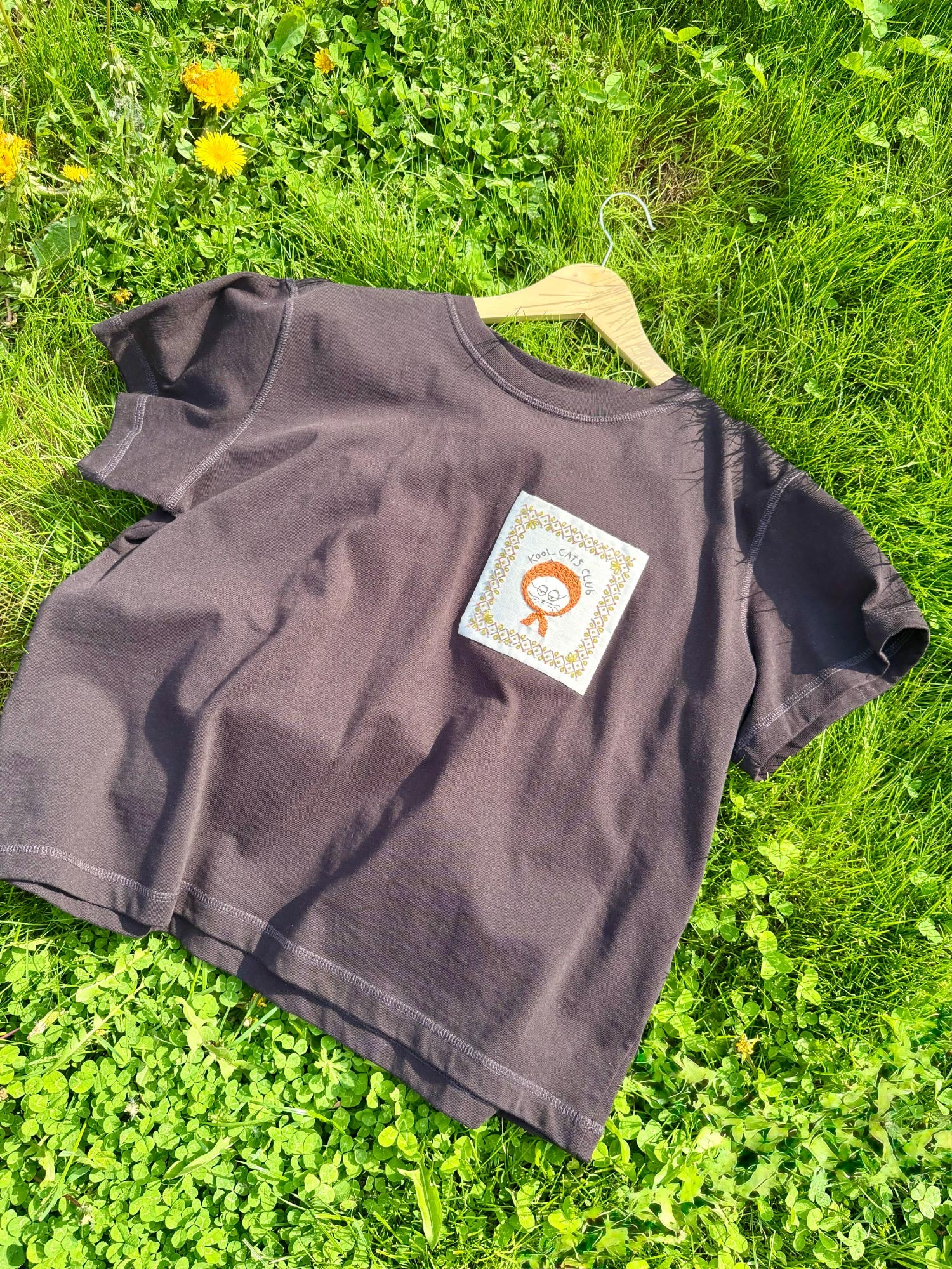 Dark purple T-shirt featuring a hand-embroidered cat design, displayed on a wooden hanger and photographed outdoors on a sunny day among the grass.