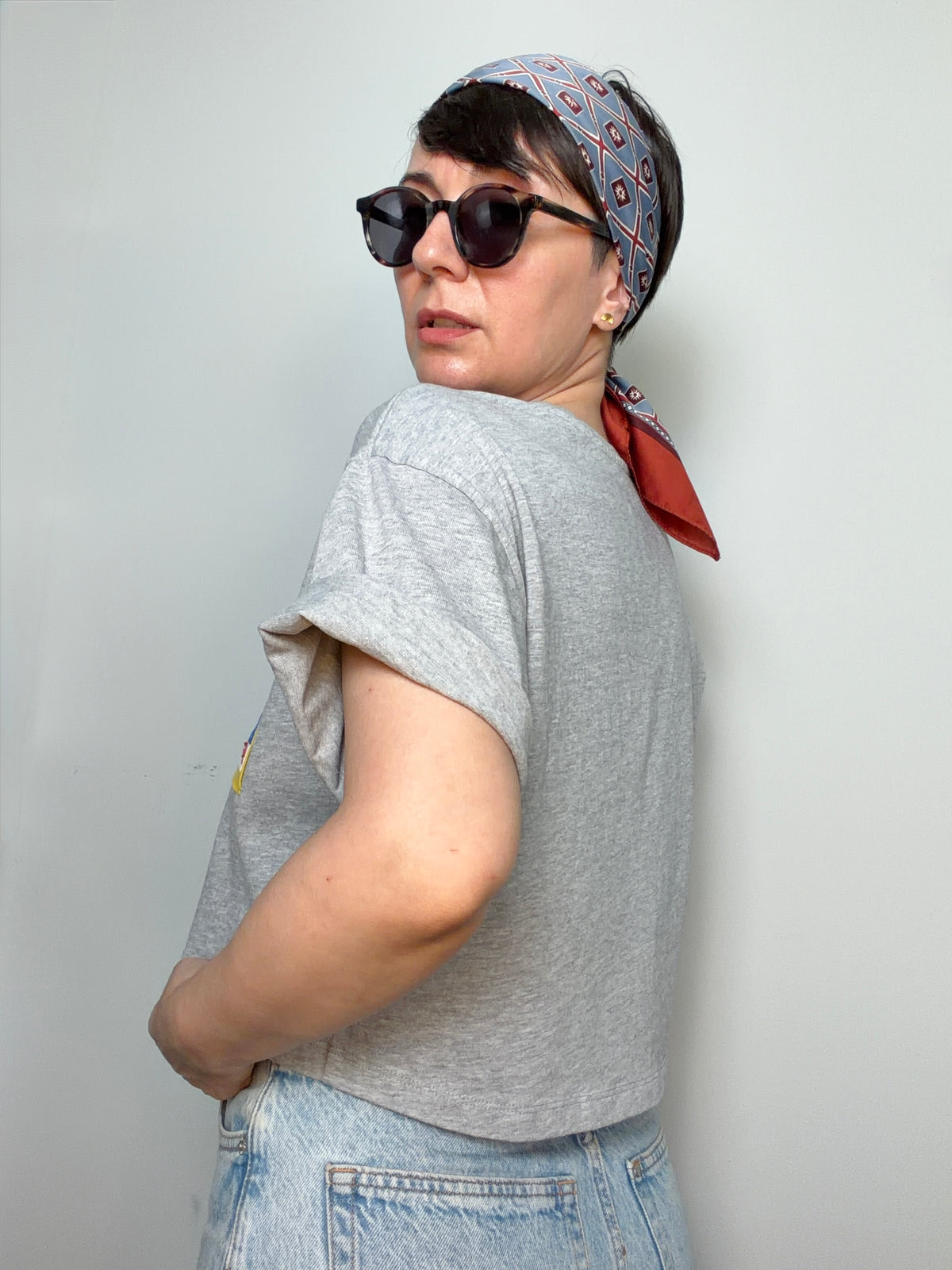 Person wearing a grey t-shirt, sunglasses, and a patterned headscarf against a plain background