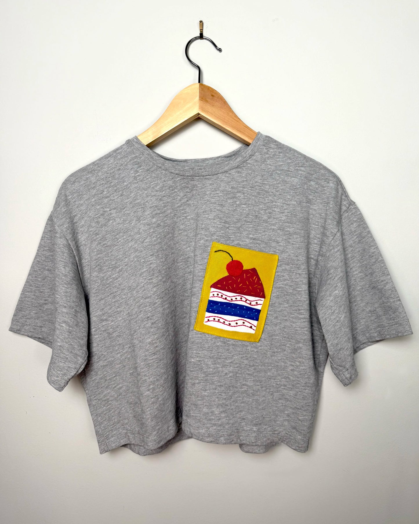 Grey t-shirt with a colourful cake appliqué on a wooden hanger against a plain background.