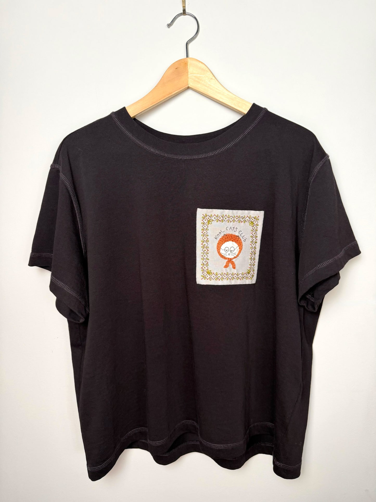 Dark purple t-shirt with hand embroidered cat design on a wooden hanger against a light grey background