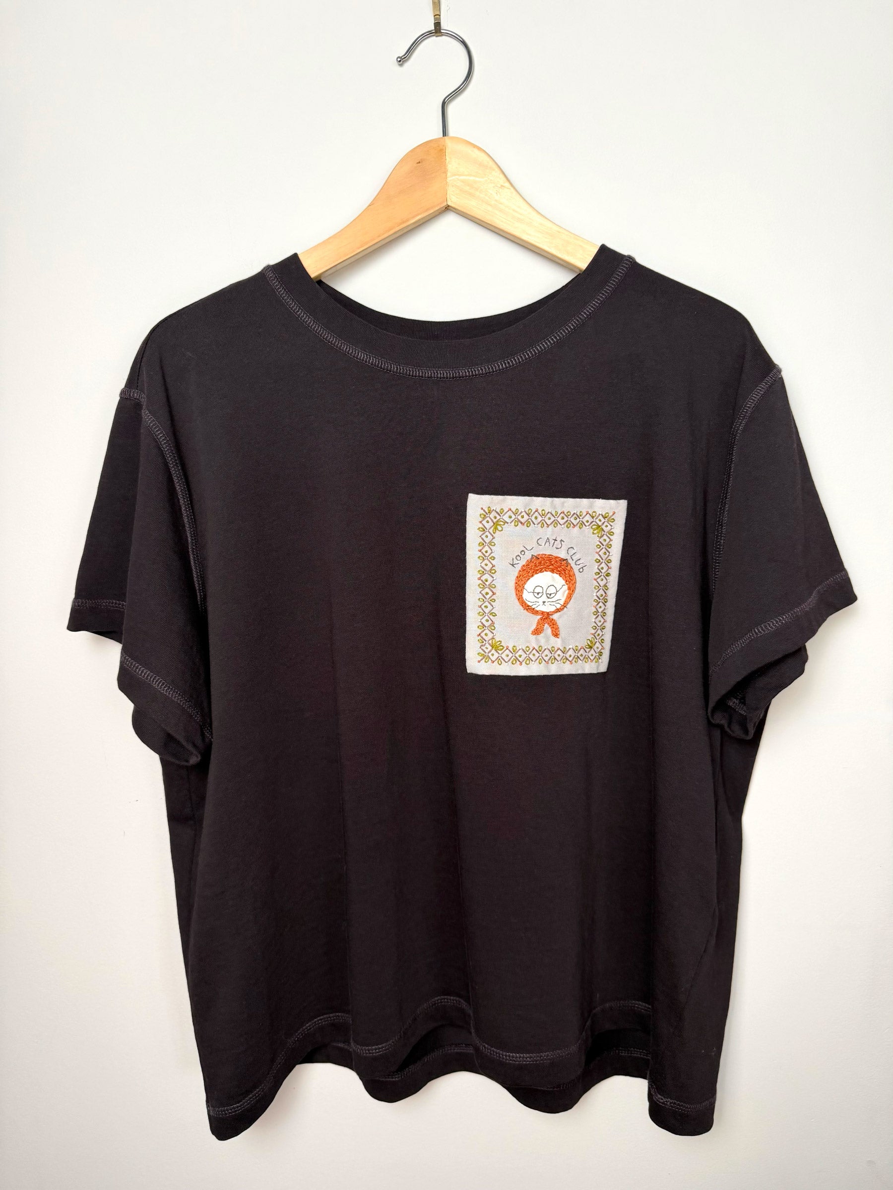 Dark purple t-shirt with hand embroidered cat design on a wooden hanger against a light grey background