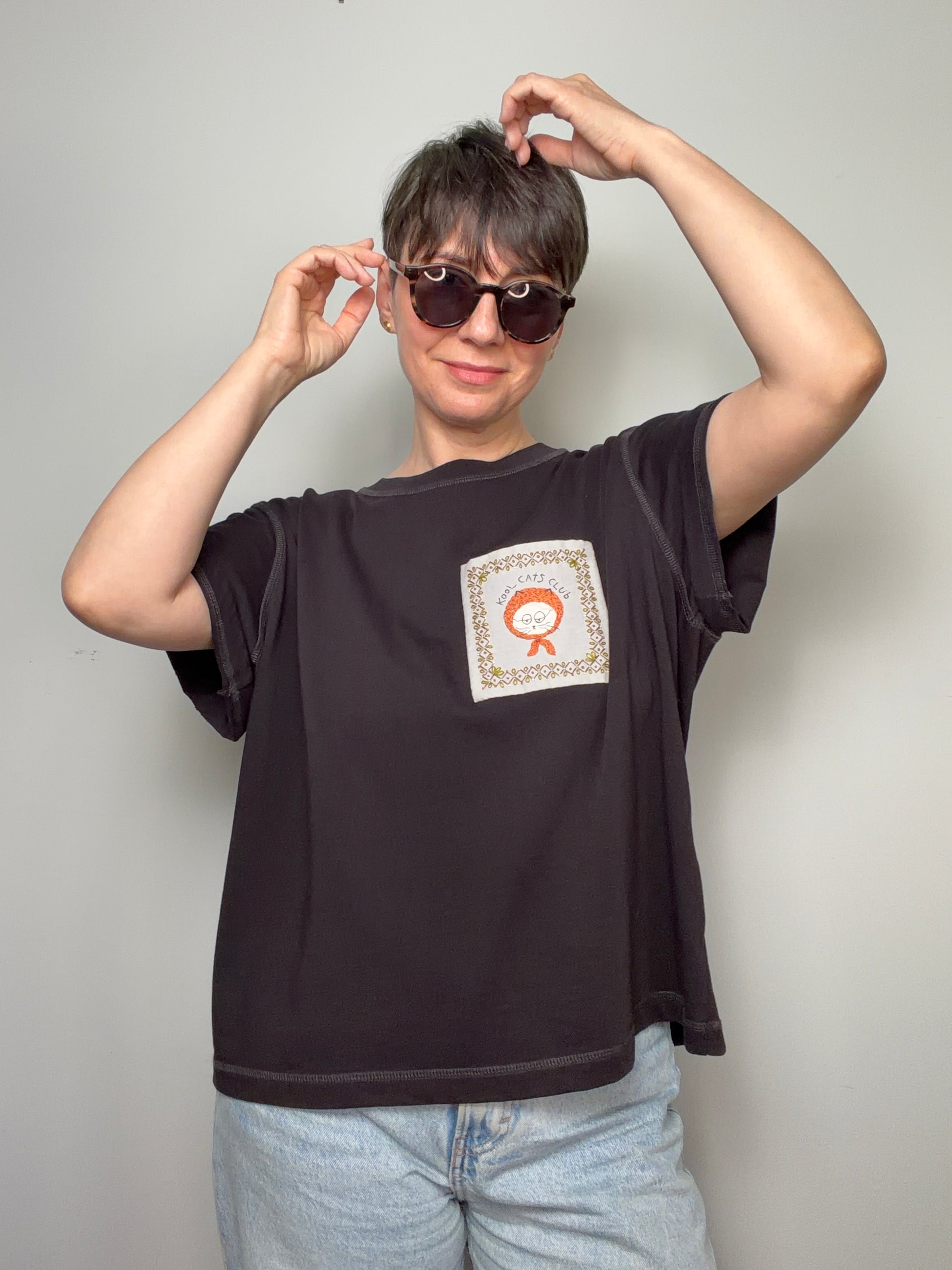 Person wearing a dark purple t-shirt with hand embroidered cat design, posing against a plain background.