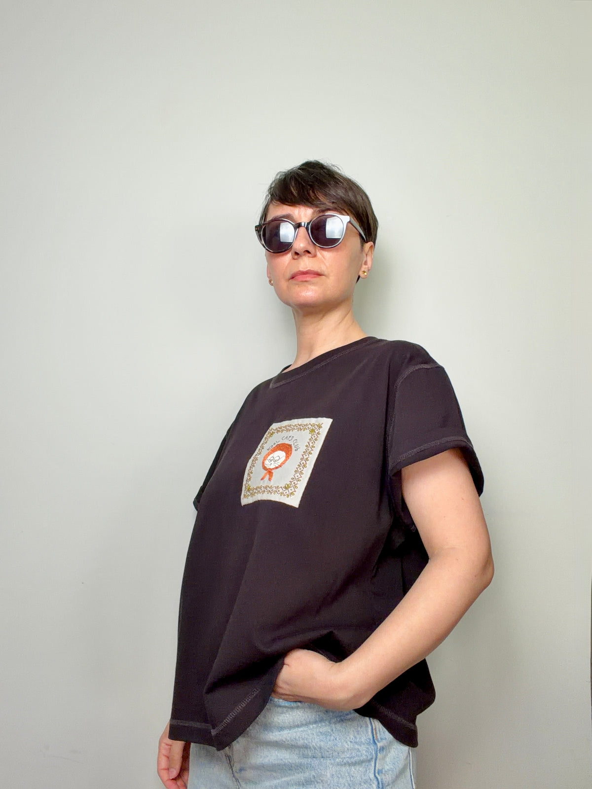 Person wearing a dark purple t-shirt with hand embroidered cat design standing in front of a plain light coloured background.