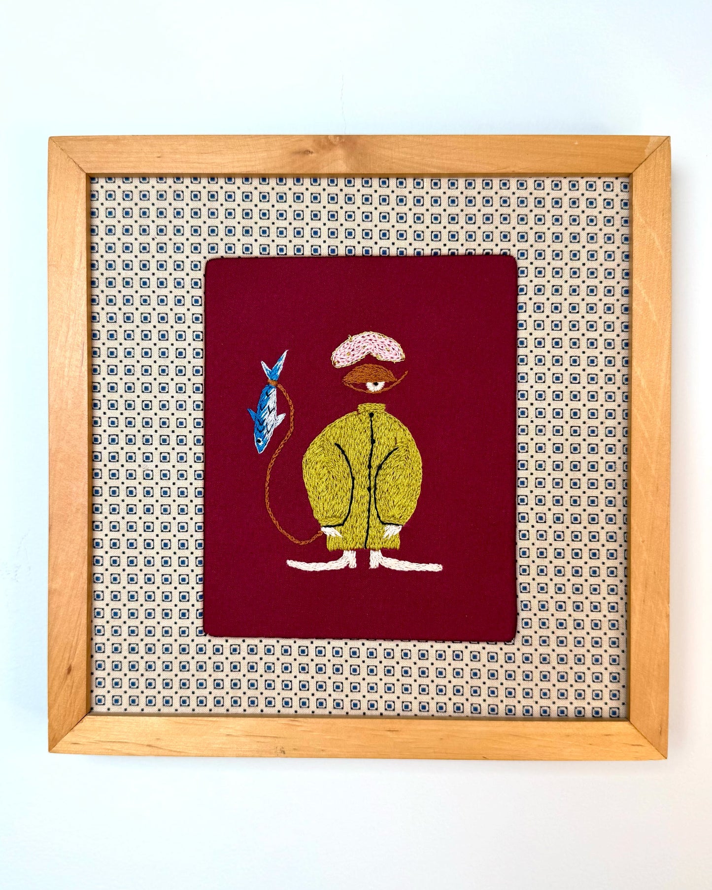 Embroidery of a character with a fish on a red background, assembled in a wood frame.